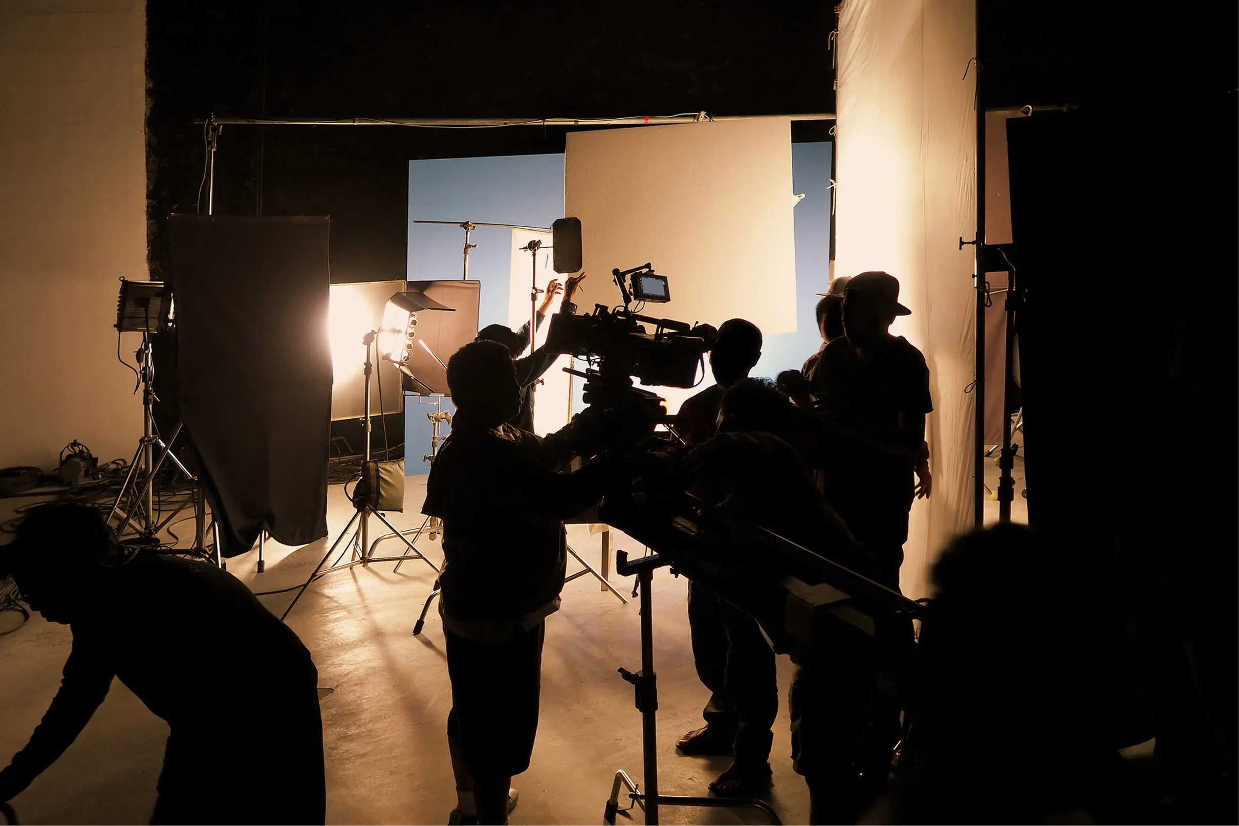GettyImages-489362405 5 (1) Film crew providing top-notch video production services work with cameras and lighting equipment on a set. Silhouetted figures adjust gear against illuminated screens and lights, ensuring every shot captures the essence of creativity in a dynamic studio environment.